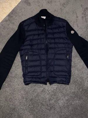 Mörkblå dunjacka från Moncler - Snygg mörkblå dunjacka från Moncler med quiltad front och stickade ärmar. Jackan har två dragkedjefickor och Moncler-logga på ena ärmen. Perfekt för kalla dagar och riktigt stilren look.