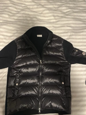 Svart pufferjacka från Moncler - Snygg svart pufferjacka från Moncler med avtagbar huva och med glansig vadderad kropp, stickade ärmar och huva. Jackan har dragkedja framtill, fickor med tryckknappar och Moncler-logga på ärmen. Perfekt för kalla vinterdagar.