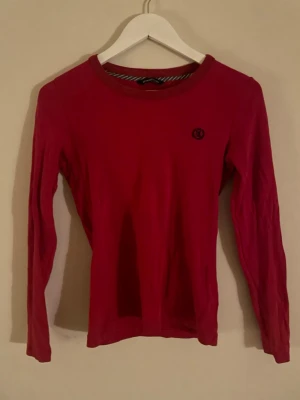 Röd långärmad topp från Henri Lloyd - Snygg röd långärmad topp från Henri-Lloyd med rund halsringning och diskret broderad logga på bröstet. Perfekt att styla med jeans eller kjol för en clean look.