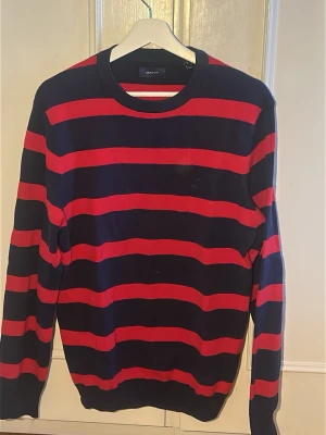 Randig tröja från Gant i marinblått/rött - Snygg långärmad tröja från Gant med marinblå bas och röda horisontella ränder. Rund halsringning och ribbade muddar vid ärmslut och nederkant. Perfekt för en avslappnad men stilren look.