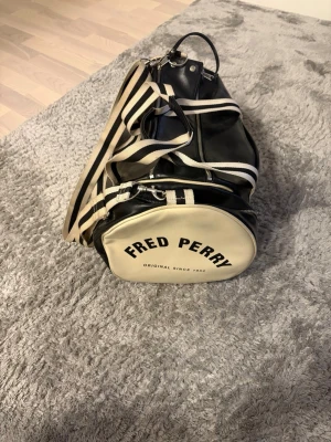 Fred Perry svart och vit väska - Snygg shoppingväska från Fred Perry i svart och vitt konstläder med klassiska ränder och logga på sidan. Väskan har cylinderform, justerbar axelrem och dragkedja. Perfekt för dig som vill ha en sportig och retro look.