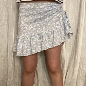 Blommig volangkjol från Gina Tricot - Supersöt kort kjol från Gina Tricot i ljusblått med vitt blommönster. Kjolen har volangkant och omlottdetalj med rynk på sidan. Perfekt för vår och sommar, lätt att matcha med crop tops eller t-shirts.