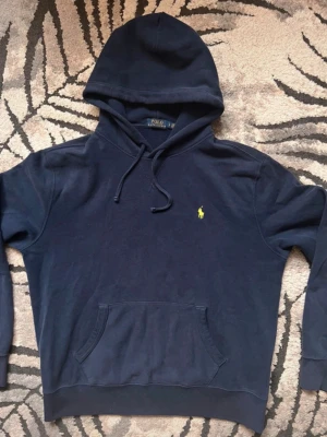 Ralph Laurent hoodie - Mörkblå hoodie från Polo Ralph Lauren med klassisk gul broderad logga på bröstet. Hoodien har huva med dragsko, känguruficka och ribbade muddar. Tillverkad i mjuk bomull för skön känsla och avslappnad passform.