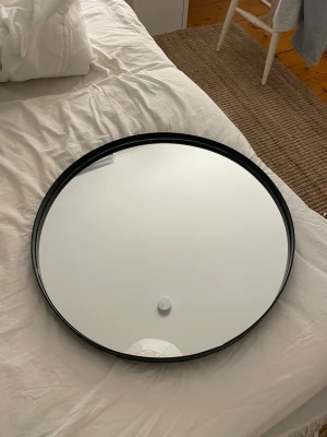 Rund spegel med svart metallram - Säljer en stilren rund spegel med svart metallram. Spegeln har en modern och enkel design som passar perfekt i hallen, sovrummet eller vardagsrummet. Baksidan är förstärkt med metallkors för extra stabilitet. Diameter ca 60 cm.