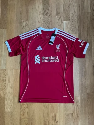 Liverpool matchtröja Adidas röd - Säljer en röd Liverpool FC matchtröja från Adidas med vita detaljer och tryck. Tröjan har klubbmärke, sponsorlogga och nummer 9 med namnet ISAK på ryggen. Perfekt för fotbollsfans som vill sticka ut på läktaren eller planen.