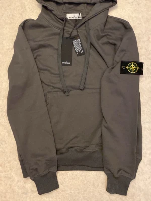 Grå hoodie från Stone Island - Snygg grå hoodie från Stone Island med klassisk logotyp-patch på vänster ärm. Hoodien har huva med dragsko, magficka och ribbade muddar. Perfekt för en avslappnad och trendig stil. Materialet är mjukt och bekvämt bomullstyg.