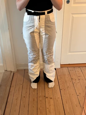Peak performance skidbyxor - Säljer ett par vita täckbyxor med svarta detaljer vid benslut och midja. Byxorna har hög midja, dragkedja fram och praktiska fickor med dragkedja på sidorna. Materialet är vadderat och passar perfekt för kalla dagar i backen. Snygg passform och stilren design.