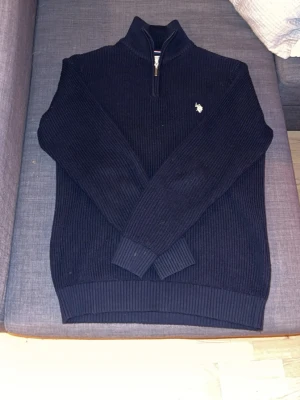 Marinblå stickad tröja U.S. Polo Assn - Mörkblå stickad tröja från U.S. Polo Assn med halv dragkedja och hög krage. Tröjan har ribbstickade muddar och en vit broderad logga på bröstet. Perfekt för dig som gillar klassisk och stilren look.