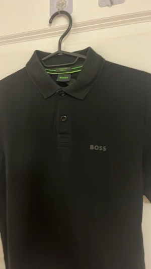 Svart pikétröja från BOSS - Äkta!!!Klassisk svart pikétröja från BOSS med korta ärmar och krage. Tröjan har två knappar framtill och diskret BOSS-logga på bröstet. Perfekt för en stilren och enkel look. Materialet känns mjukt och bekvämt.