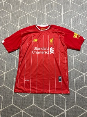 New Balance Liverpool - Säljer en röd Liverpool FC matchtröja från New Balance med vita tunna ränder, klubbmärke och sponsortryck på bröstet. Tröjan har rund hals med vit kant och gula detaljer. Perfekt för dig som älskar fotboll och vill stötta ditt lag.