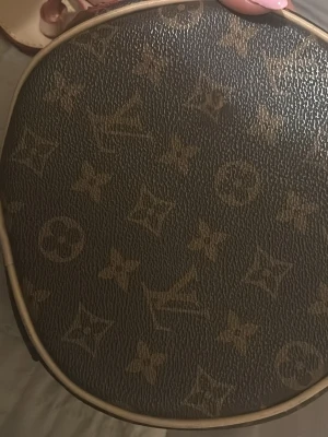 Louis Vuitton rund monogramväska - Säljer en rund handväska från Louis Vuitton med klassiskt brunt monogrammönster och beigea detaljer. Väskan har en exklusiv LV-tag i guld och beige läder. Perfekt accessoar för dig som gillar lyxiga och tidlösa designer. AAA KOPIA