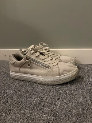 Beige sneaker från Graceland!! - Stilrena beige sneakers i mockaimitation med snörning och en snygg silverfärgad dragkedja på sidan. Skorna har en vit, räfflad sula och diskreta detaljer vid hälen. Perfekta för en avslappnad och trendig look.