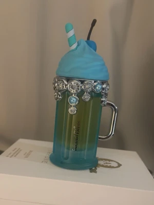 Island Bliss parfym milkshake-flaska - Unik parfymflaska från Island Bliss formad som en milkshake i blått och grönt glas. Locket är dekorerat med blått lock, sugrör och körsbär samt gnistrande stenar och pärlor runt kanten. Handtag i silverfärg ger extra cool detalj. Perfekt för dig som gillar lekfull design.