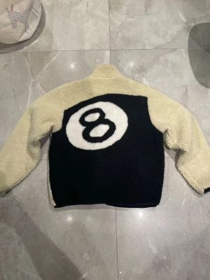 Stussy teddyjacka med 🎱-ball  - Säljer en beige teddyjacka från Stussy med svart bröstficka och dragkedja. Baksidan har en stor svart panel med en vit 8-ball. Jackan är fluffig och har hög krage samt detaljer i svart. Perfekt för dig som gillar streetwear och unika plagg.  Fåtal gånger använd och använt sparsamt. Inga defekter storlek S med cropped midja