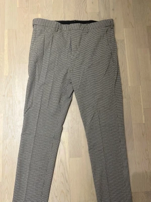 Rutiga kostymbyxor i svartvitt - Snygga kostymbyxor med svartvitt rutmönster och raka ben. Byxorna har bälteshällor, sidofickor och klassisk passform. Perfekta för dig som vill ha en stilren och trendig look. Materialet känns lätt och passar året runt.