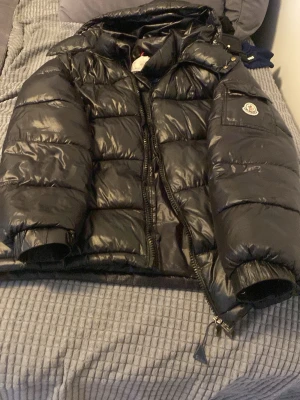 Svart dunjacka från Moncler - Säljer en svart, glansig dunjacka från Moncler med klassisk logotyp på ärmen och praktisk ficka med knapp. Jackan har hög krage, dragkedja och breda kanaler fyllda med dun för extra värme. Perfekt för kalla vinterdagar.