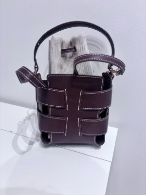 Bucketbag - Säljer en unik brun axelväska i flätad läderlook med vita kontrastsömmar och justerbar axelrem. Väskan har en innerpåse i beige canvas med dragsko, vilket ger en cool och modern vibe. Perfekt för dig som gillar stilrena detaljer och smart förvaring.