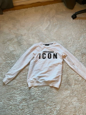 Vit sweatshirt DSQUARED2 ICON - Säljer en vit sweatshirt från DSQUARED2 med svart text 'ICON' och logga framtill. Tröjan har rund halsringning, långa ärmar och ribbade muddar. Materialet är mjukt och bekvämt, perfekt för en avslappnad stil.sitter som CS