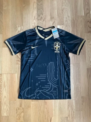 Svart Brasilien Nike matchtröja - Snygg svart Brasilien matchtröja från Nike med gulddetaljer och grafiskt mönster framtill. Tröjan har korta ärmar, V-ringning och Brasilien-emblem på bröstet. Tillverkad i lätt och ventilerande funktionsmaterial, perfekt för fotboll eller träning.