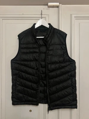 Svart dunväst med ståkrage - Snygg svart dunväst med ståkrage och quiltad design. Västen har dragkedja framtill och ett stilrent, minimalistiskt utseende. Perfekt att slänga över en hoodie eller tröja för extra värme. Tillverkad i lätt och smidigt material som håller dig varm utan att kännas klumpig.