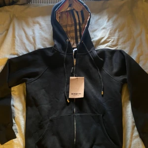 Svart Burberry hoodie med dragkedja - Svart hoodie från Burberry med klassiskt rutigt foder i huvan. Jackan har dragkedja framtill, snörning i huvan och två fickor. Tillverkad i mjuk bomull och har en stilren, tidlös look med exklusiva detaljer. Det står att den är L fast den passar M
