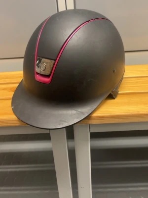 Svart ridhjälm från Samshield - Snygg svart ridhjälm från Samshield med en matt finish och rosa detalj framtill. Hjälmen har en klassisk form med skärm och ventilationshål på sidorna. Insidan är vadderad för extra komfort och har ett justerbart hakband.