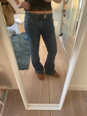 Blå bootcut jeans med låg midja - Säljer ett par blå bootcut jeans med låg midja och dubbla knappar i linningen. Jeansen har klassiska fem fickor och är tillverkade i ett mjukt denimtyg. Perfekta för en avslappnad och trendig look. En fläck på ena fickan då jag hade ett trasigt läppglans i fickan. Har aldrig försökt få bort det så vet inte om det går att få bort i tvätten eller inte🥰🥰