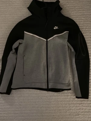 Nike svart och grå helt ny äkta  - Nike vindjacka i svart och grått med hel dragkedja och hög krage. Jackan har vit logga på bröstet, snyggt mönster över bröstet och praktiska fickor. Perfekt för blåsiga dagar och sportiga outfits.