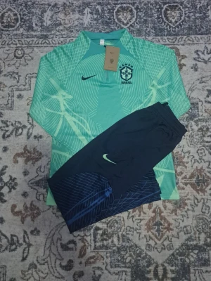 Nike Brasil träningsset mintgrön/blå - Säljer ett Nike Brasil träningsset med mintgrön långärmad tröja och marinblå byxor. Tröjan har half zip, Brasil-logga och ett grafiskt mönster. Byxorna är mörkblå med ljusgröna detaljer och Nike-logga. Perfekt för fotbollsträning.BARNSIZE 18(XL BARN)