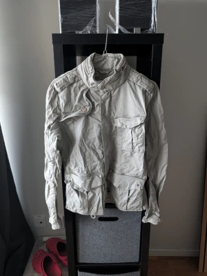 Beige utilityjacka med fickor - Snygg beige utilityjacka med flera stora fickor framtill, hög krage och knappar vid ärmslut. Jackan har en avslappnad passform och är tillverkad i bomull. Perfekt för dig som gillar en stilren och funktionell look.