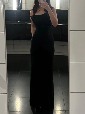 Svart axelbandsklänning i sammet - Elegant lång svart klänning i sammet med asymmetrisk axel och figurnära passform. Klänningen har en avskuren axel och en mjuk, stretchig känsla som framhäver siluetten. Perfekt för dig som vill sticka ut med en stilren look.