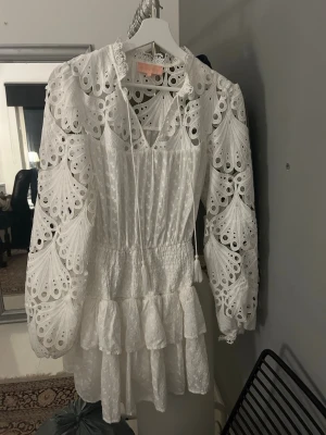 Feeling Friday Vit kortklänning med volang - Superfin vit kortklänning från Feeling Friday.  Broderade detaljer och volanger. Klänningen har långärmade puffärmar med hålmönster, v-ringning och smock i midjan för en snygg passform. Perfekt för sommarens alla tillfällen och tex studenten! Har använts bara en gång, den är som ny!