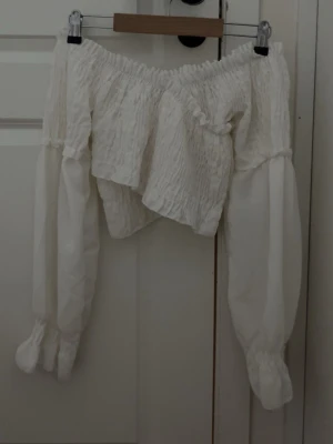 Vit offshoulder blus med puffärm - Trendig vit offshoulder blus med smockad överdel och långa puffärmar. Blusen har resår vid axlar och ärmslut för en snygg passform och romantisk känsla. Perfekt att styla med jeans eller kjol för en cool look.