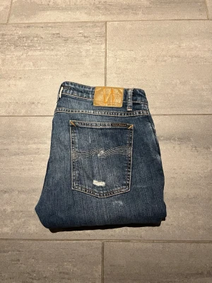 Blå slitna jeans från Nudie Jeans - Säljer ett par blå jeans från Nudie Jeans med skinny fit och slitningar på både fram- och baksida. Klassisk femficksmodell med läderpatch bak i midjan och snygga detaljerade sömmar på bakfickan. Tillverkade i denim av bomull.