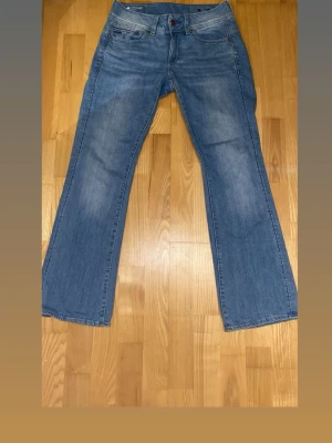 Blå utsvängda jeans från G-Star - Säljer ett par blå lågmidjade jeans från G-Star, modell Midge. Jeansen har utsvängda ben. Dem är storlek 27 men passar även 26 då dem har en knapp i midjan bak (se bild 4). Längd 30