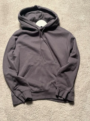 Mörkgrå basic hoodie med huva - En stilren mörkgrå hoodie med huva och dragsnören. Hoodien har en klassisk känguruficka framtill och ribbade muddar vid ärmar och nederkant. Perfekt för en avslappnad och trendig look. Tillverkad i mjukt bomullsmaterial.