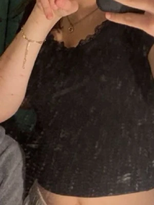 Svart spetskantad croppad topp - Snygg svart croppad topp med spetskant vid urringningen. Perfekt att styla med höga jeans eller kjol för en trendig look. Toppen är ärmlös och har en feminin touch tack vare spetsdetaljen.