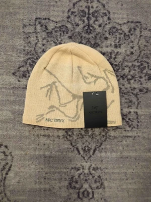 Arc'teryx beige mössa i ull - Snygg beige mössa från Arc'teryx med grått grafiskt mönster och broderad logga framtill. Tillverkad i mjukt stickat material som sitter skönt på huvudet. Perfekt för dig som vill ha en stilren och modern accessoar till höst och vinter.