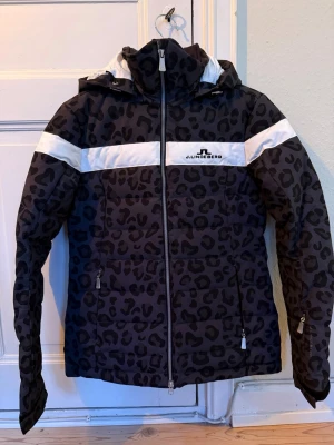 Svart leopardmönstrad skidjacka J.Lindeberg - Svart/grå fodrad skidjacka från J.Lindeberg med leopardmönster och vita detaljer över bröstet och axlarna. Bara använd 2 veckor. Jackan har huva, hel dragkedja och två fickor med dragkedja samt en innerficka och ficka i ärmen för liftkortet. Perfekt för dig som vill sticka ut i backen! Nypris ca 5000 