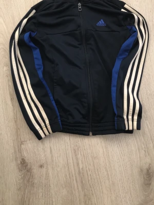 Svart Adidas träningsjacka 140 - Snygg svart träningsjacka från Adidas med dragkedja, vita klassiska ränder längs ärmarna och blå detaljer. Perfekt för träning eller chill. Tillverkad i ett lätt och bekvämt material som andas. Adidas-logga på bröstet.