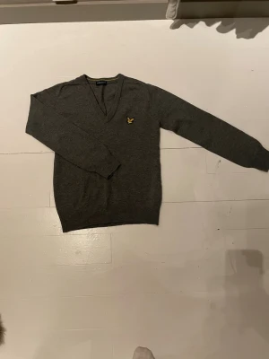 Grå stickad tröja från Lyle & Scott - Mörkgrå stickad tröja med V-ringning från Lyle & Scott. Tröjan har lång ärm och den klassiska gula loggan broderad på bröstet. Perfekt för lager på lager och enkel att matcha med olika outfits.