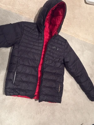 Svart dunjacka med röd insida från BOSS - Snygg svart dunjacka från BOSS med röd insida och huva. Jackan har två dragkedjeförsedda fickor och BOSS-logga på bröstet. Perfekt för kalla vinterdagar och riktigt skön att bära tack vare det vadderade materialet. går att ha ut och in. 14 år/s