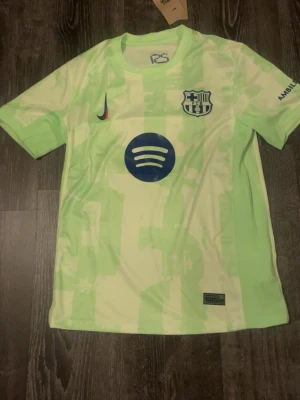 Barça matchtröja ljusgrön Nike - Säljer en ljusgrön FC Barcelona matchtröja från Nike med blå loggor och tryck, inklusive Spotify och UNHCR. Tröjan har korta ärmar, rund hals och modern design med mönstrad yta. Perfekt för dig som älskar fotboll och vill sticka ut på planen.