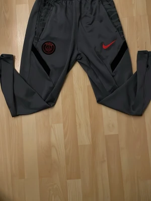 Gråa Nike PSG träningsbyxor - Snygga gråa träningsbyxor från Nike med PSG-logga på vänster ben och röd Nike-swoosh på höger ben. Byxorna har svarta detaljer längs sidorna och är tillverkade i ett mjukt, stretchigt material som passar perfekt för sport och träning.
