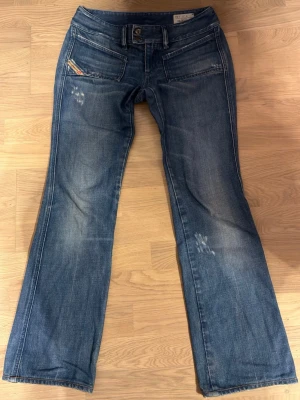 Blå bootcut jeans med extremt lågmidja från Diesel - Säljer ett par blå bootcut jeans från Diesel med slitningar och tvättade detaljer. Jeansen har dubbla knappar i midjan, stora fickor fram och en liten logga på fickan. Klassisk denimlook med coola detaljer och avslappnad passform. De är extremt lågmidjade och bootcut. Dessa är även i lerfekt skick och har inga defekter då de aldrig har kommit till nån användning och säljer darför dessa!