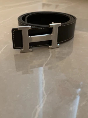 Silver skärp med H-spänne - Snyggt svart skärp med det ikoniska H-spännet i silverfärgad metall. Bältet har vita kontrastsömmar längs kanterna och är gjort i slätt skinn. Perfekt accessoar för att lyfta vilken outfit som helst.