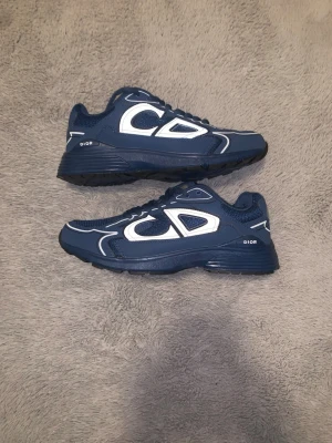 Blå Dior sneakers med vita detaljer - Snygga blå sneakers från Dior med vita detaljer och CD-logga på sidan och hälen. Skorna har en sportig design med mesh och syntetmaterial, samt robust sula för extra komfort. Perfekta för dig som vill sticka ut med exklusiva sneakers.