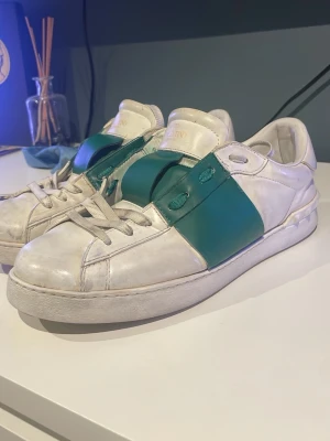 Valentino vita sneakers med grön detalj - Säljer ett par vita sneakers från Valentino med en bred grön läderdetalj över snörningen. Skorna har klassisk låg profil, vita snören och små nitar på hälen. Perfekta för dig som vill ha något unikt och stilrent.
