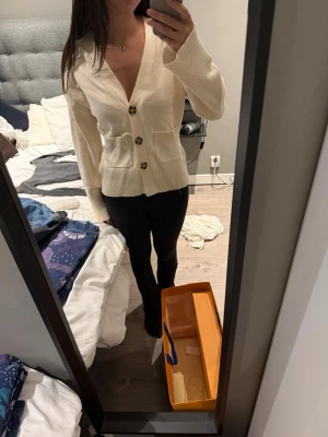 Krämvit kofta med stora knappar - Snygg krämvit kofta med djup V-ringning och tre stora, mönstrade knappar framtill. Koftan har långa ärmar, ribbstickade muddar och två fickor framtill. Perfekt för lager-på-lager och enkel att matcha med olika outfits.