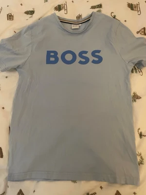 Ljusblå BOSS t-shirt med tryck - Snygg ljusblå t-shirt från BOSS med stort blått BOSS-tryck på bröstet. Klassisk rund hals och korta ärmar. Tillverkad i mjuk bomull som känns skön mot huden. Perfekt för en clean och avslappnad stil.